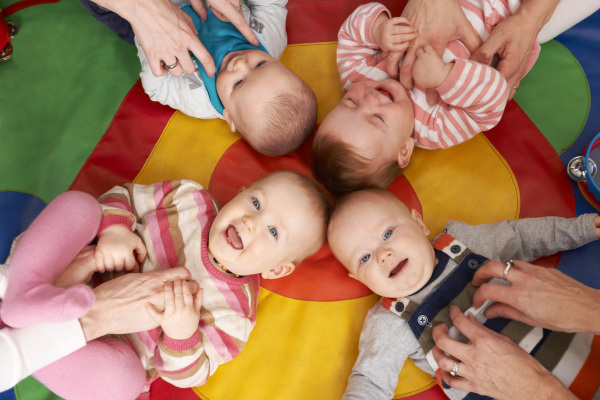 Vier Babys liegen Kopf an Kopf auf einer Krabbeldecke, werden gekitzelt und lachen.